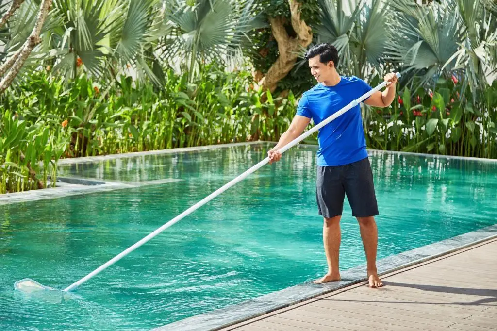Smiling man in blue shirt and black shorts cleaning a swimming pool
